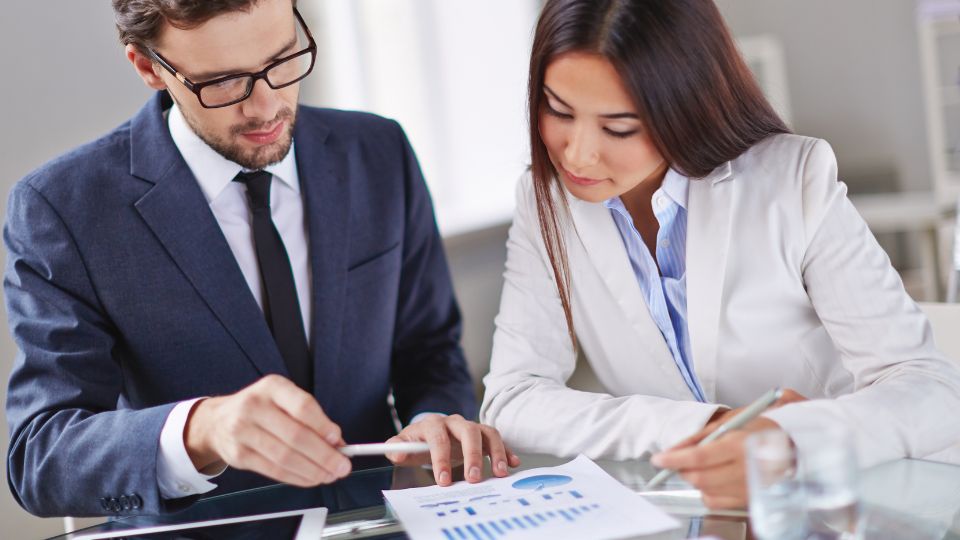 Two people sitting at a desk and working on a document with various metrics on it.