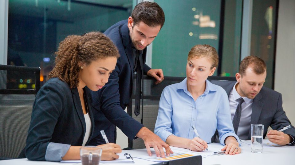 Four business professionals are reviewing documents and writing notes around a conference table in a meeting.